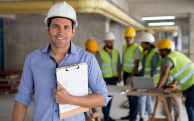 Portrait of cheerful architect at a construction site holding a clipboard smiling at camera - Incidental people working at background